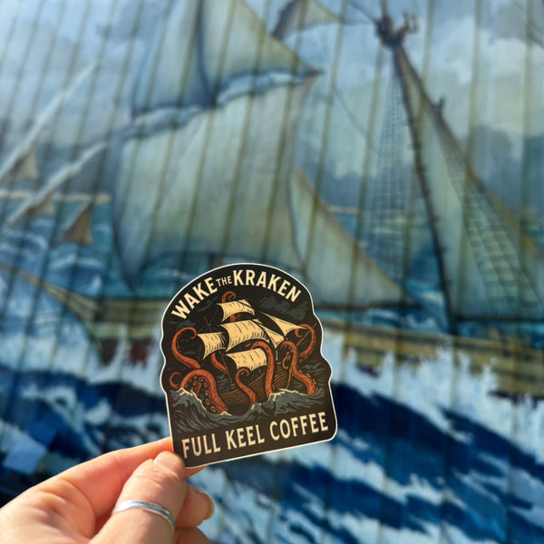 Sticker with 'Wake the Kraken Full Keel Coffee' design held in front of a ship painting.