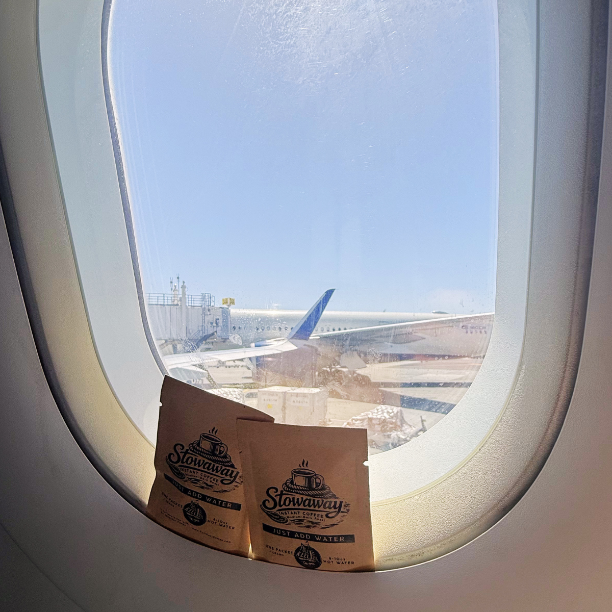 Two Stowaway instant specialty coffee packets in front of an airplane window with a view of another aircraft on the tarmac.