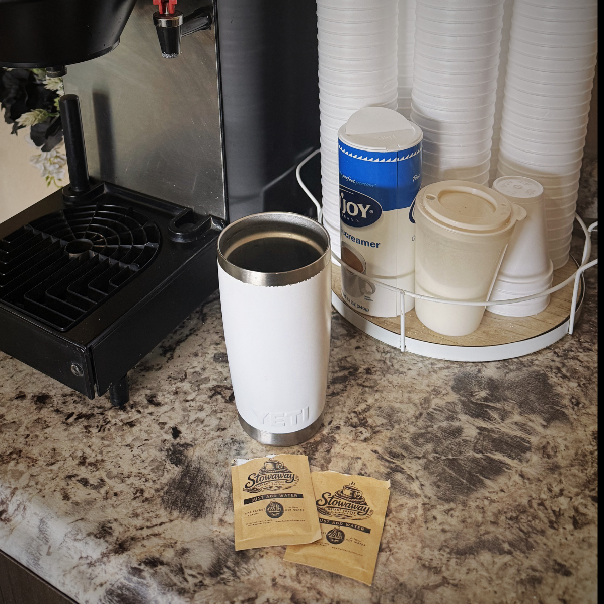 Coffee machine with a white tumbler, stowaway instant coffee packets, and disposable cups on a countertop.