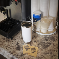 Coffee machine with a white tumbler, stowaway instant coffee packets, and disposable cups on a countertop.